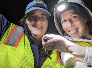 Two women wearing reflective vests and headlamps smile for a selfie with a spotted salamander. (photo © Sarah Wilson)