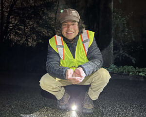 A young man wearing a reflective vest smiles while crouching down on a road and holding a wood frog in his hands. (photo © Brett Amy Thelen)