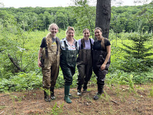 Three undergraduate interns and their mentor wearing chest waders pose for a photo at the edge of a green wetland. (photo © Brett Amy Thelen)