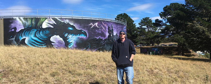 A panoramic photo of Jake Thomashow in front of one of his murals.
